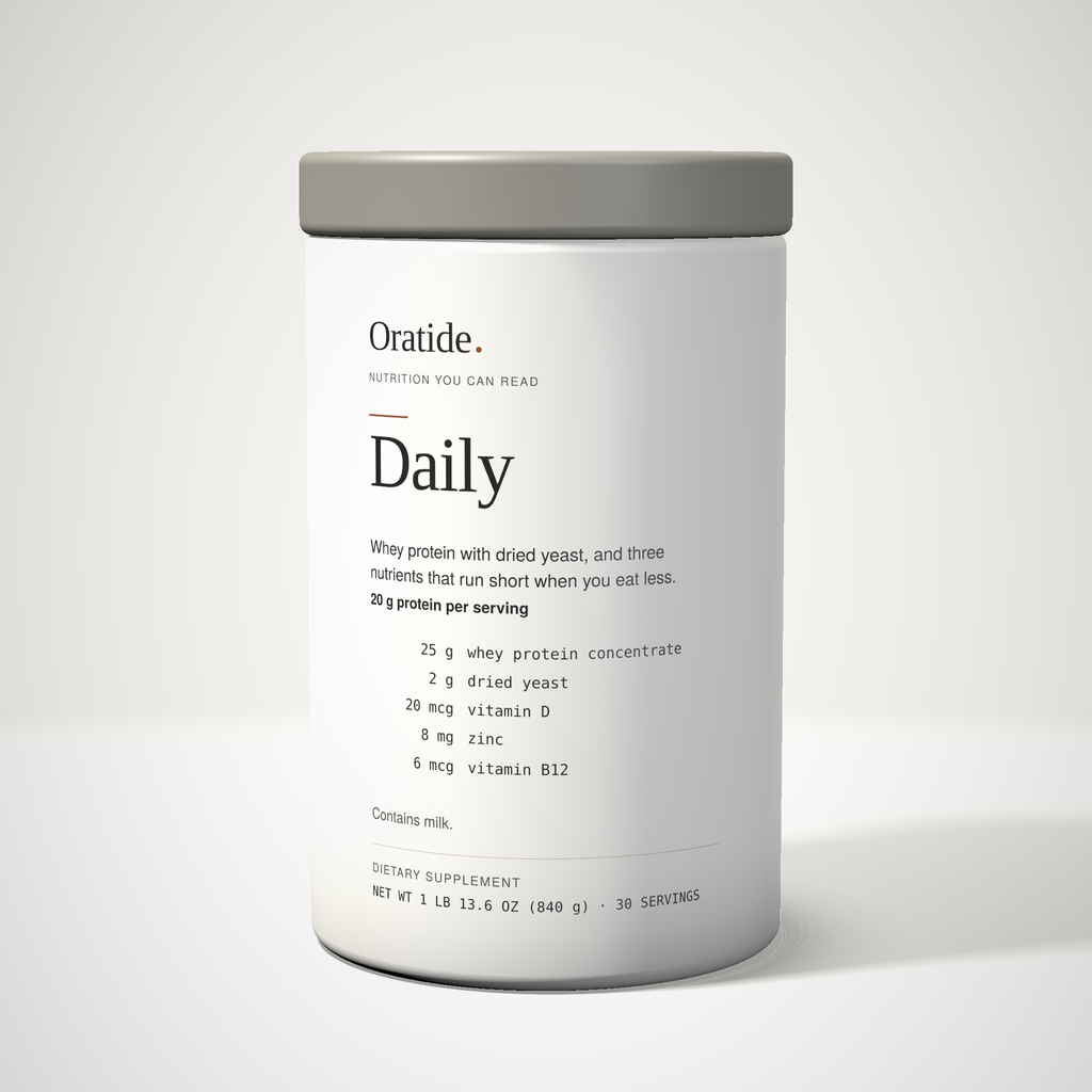 Oratide Daily, a cream 840 g canister with a grey lid, turned slightly and lit from the left on a pale surface.