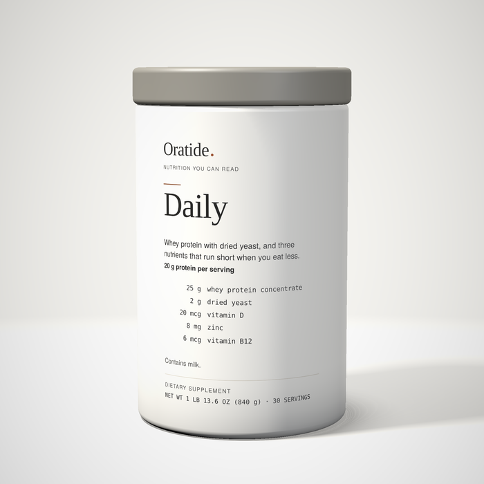 Oratide Daily, a cream 840 g canister with a grey lid, turned slightly and lit from the left on a pale surface.